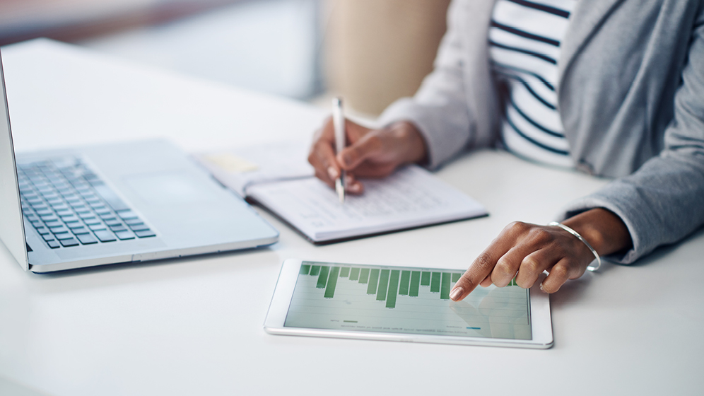 A woman writes in a notebook while reviewing data on a tablet.