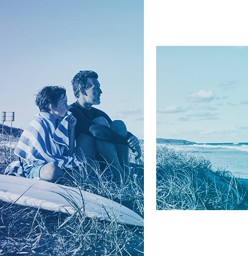 Boy and man sitting on the beach next to a surfboard, looking out at the ocean.