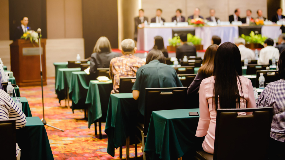 Investors sitting in rows of tables at a shareholder meeting, facing a man speaking from a podium. 